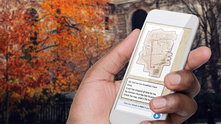 A hand holding a phone playing Treasure Hunt York with a tree with orange leaves behind.
