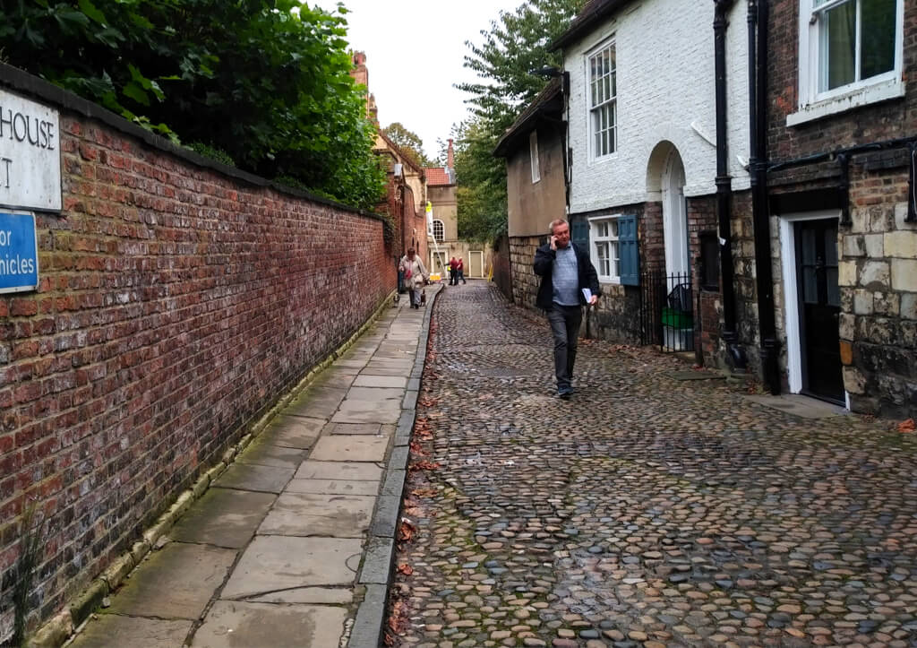 A narrow cobbled street in York, bordered by a brick wall on one side and historic buildings on the other, with a few people walking along the path.