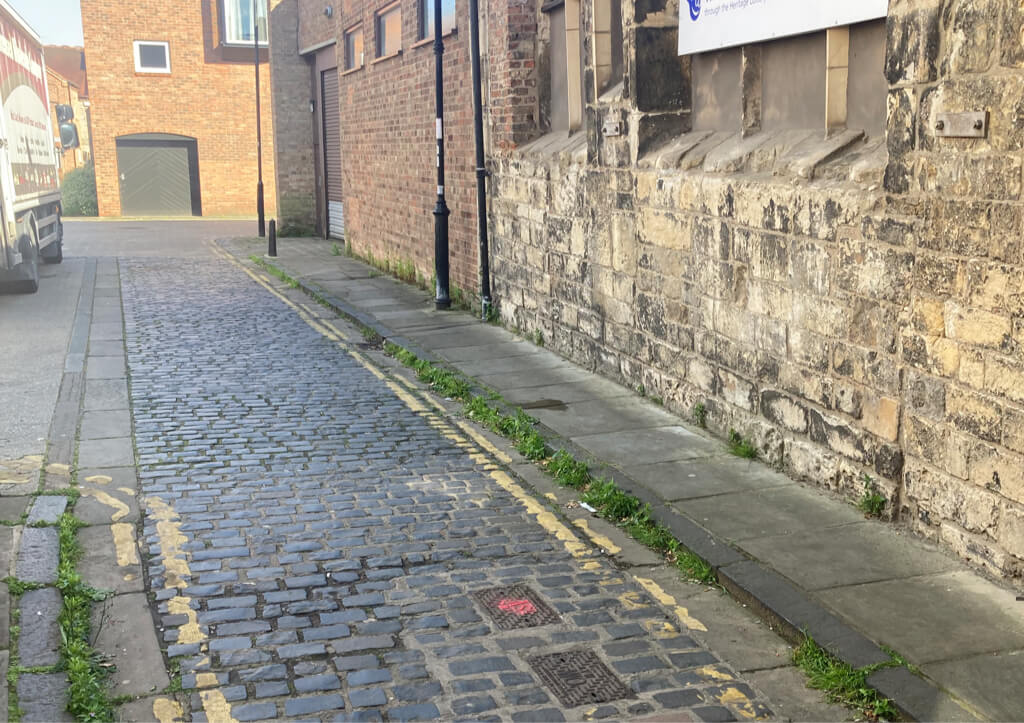 A cobbled street in York with a mix of cobblestones and paving stones, bordered by historic buildings on one side and a brick wall on the other.